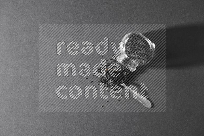 وعاء بهارات زجاجي مقلوب ممتلئ ببذور حبة البركة والبذور تناثرت منه مع ملعقة معدنية ممتلئة على أرضية سوداء