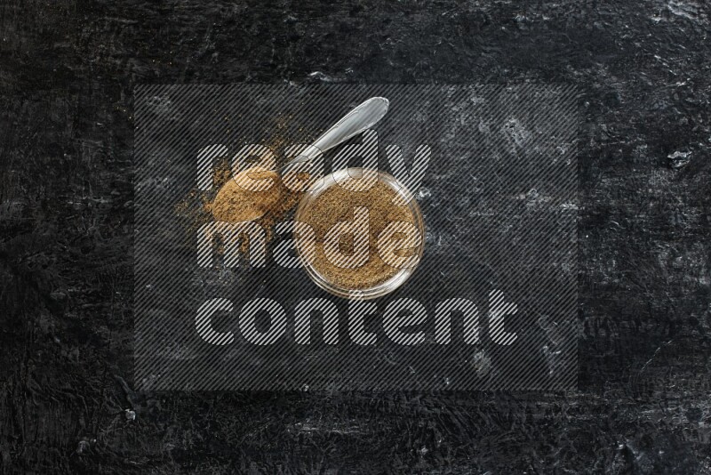 A glass jar and a metal spoon full of cumin powder on a textured black flooring