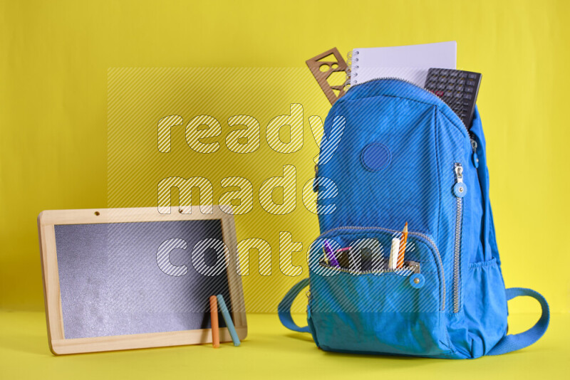 A school bag with assorted school supplies in and beside it on yellow background