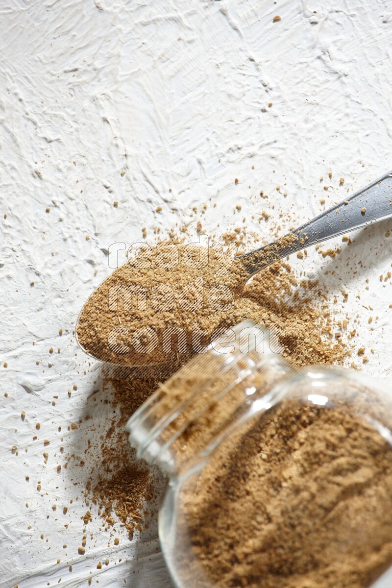 A flipped glass spice jar and a metal spoon full of cumin powder and powder spilled out on textured white flooring