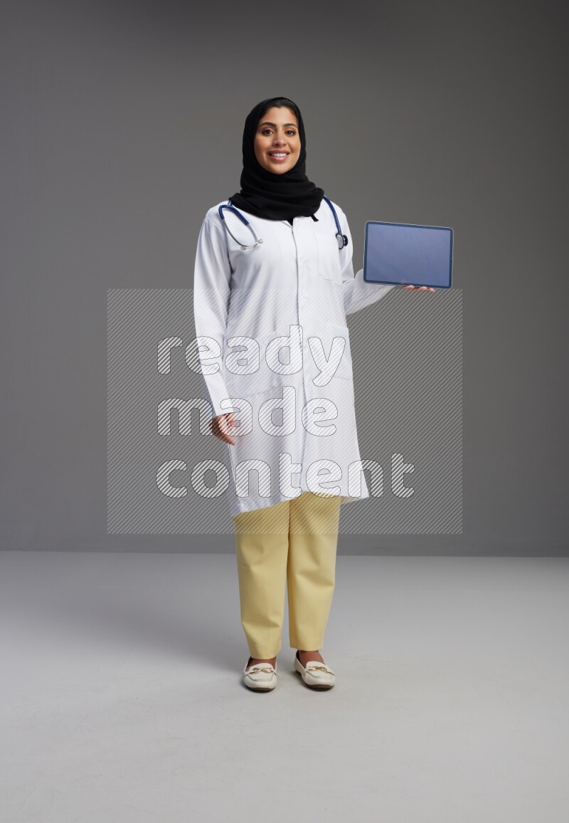 Saudi woman wearing lab coat with stethoscope standing showing tablet to camera with sign in the back on Gray background