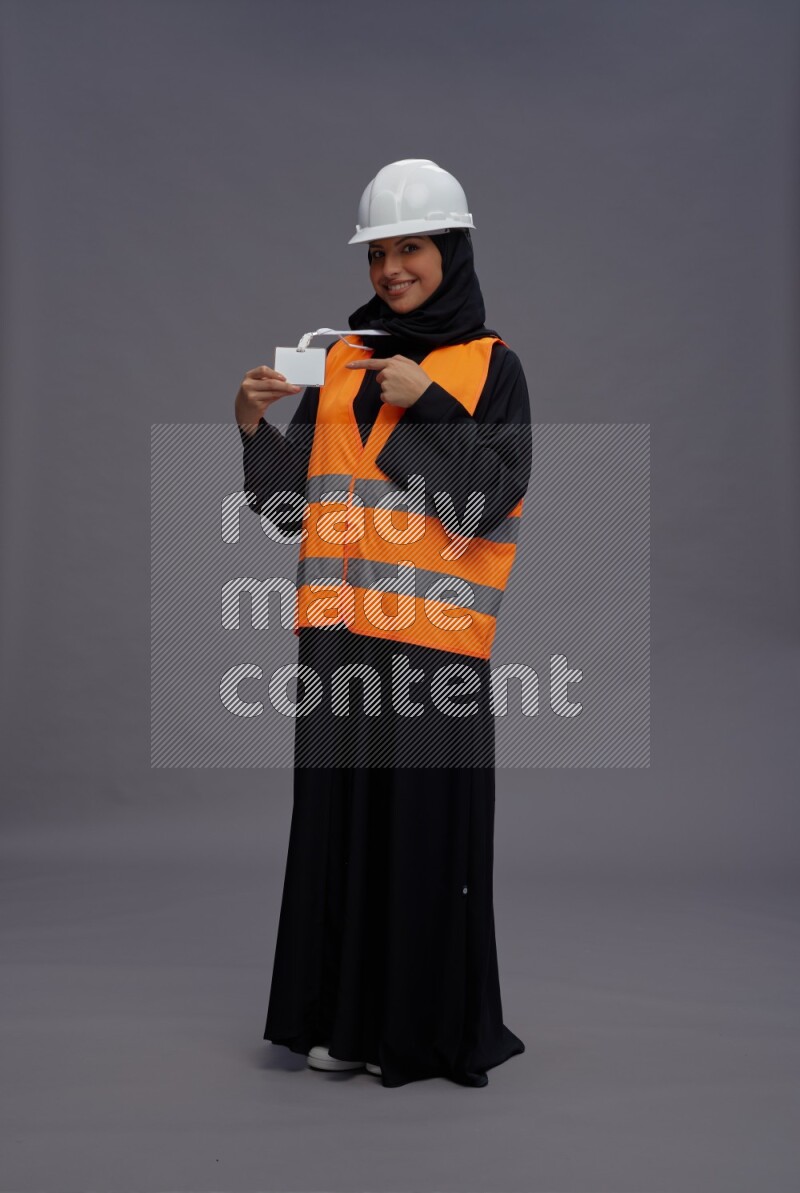 Saudi woman wearing Abaya with engineer vest with neck strap employee badge standing interacting with the camera on gray background