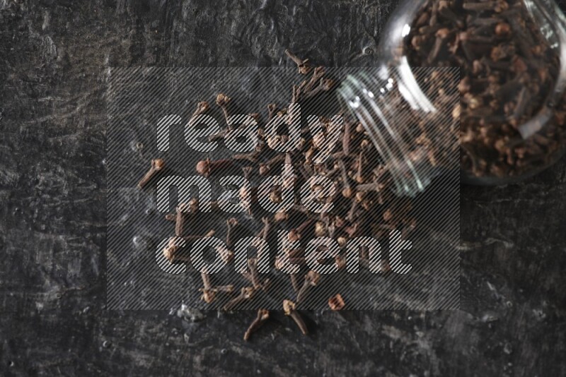 وعاء زجاجي للبهارات مقلوب ممتلئ بالقرنفل والقرنفل سقط منه على أرضية سوداء