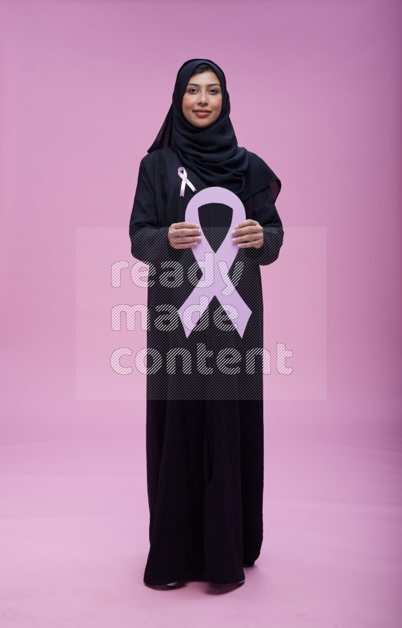 Saudi woman wearing pink ribbon on abaya standing holding awareness ribbon on plain pink background