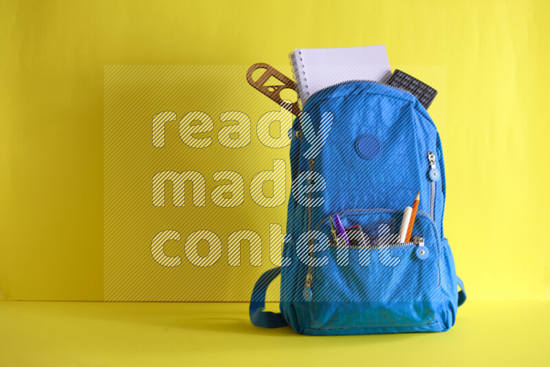 A school bag with assorted school supplies in and beside it on yellow background