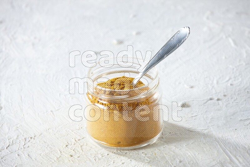 A glass jar and a metal spoon full of turmeric powder on a textured white flooring