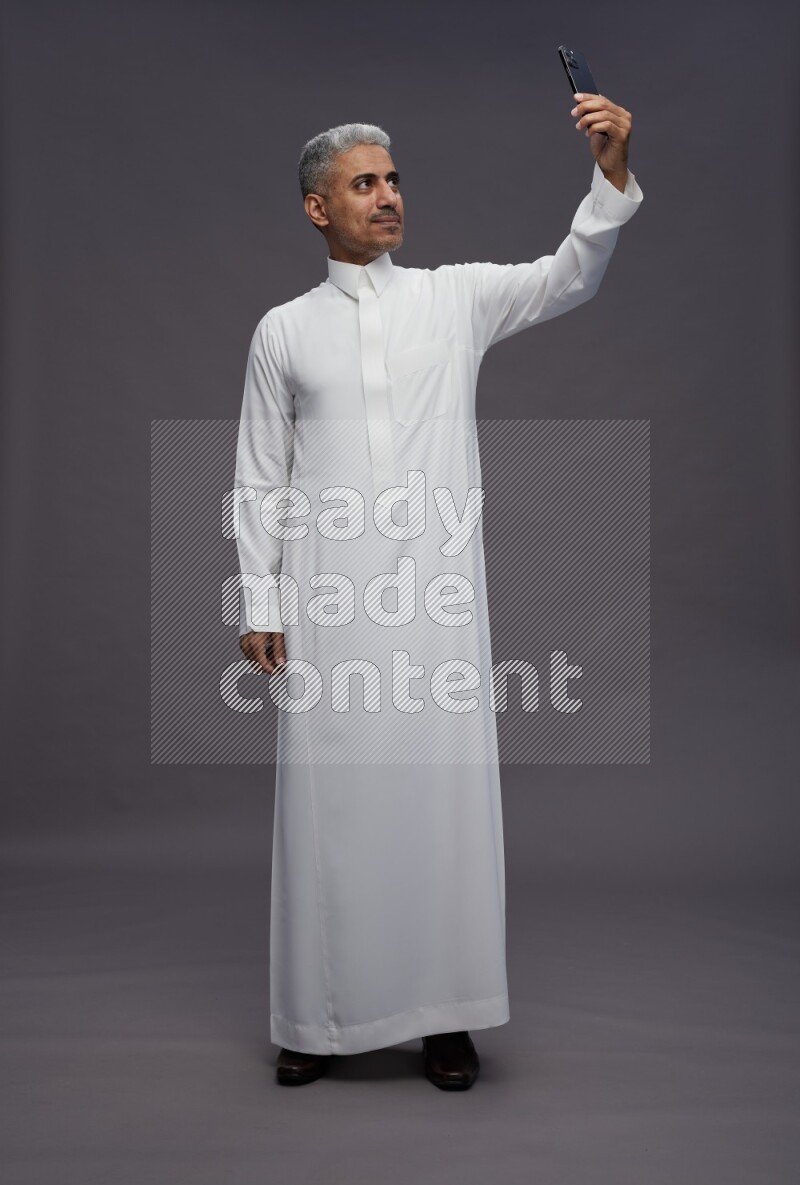 Saudi man wearing thob standing taking selfie on gray background