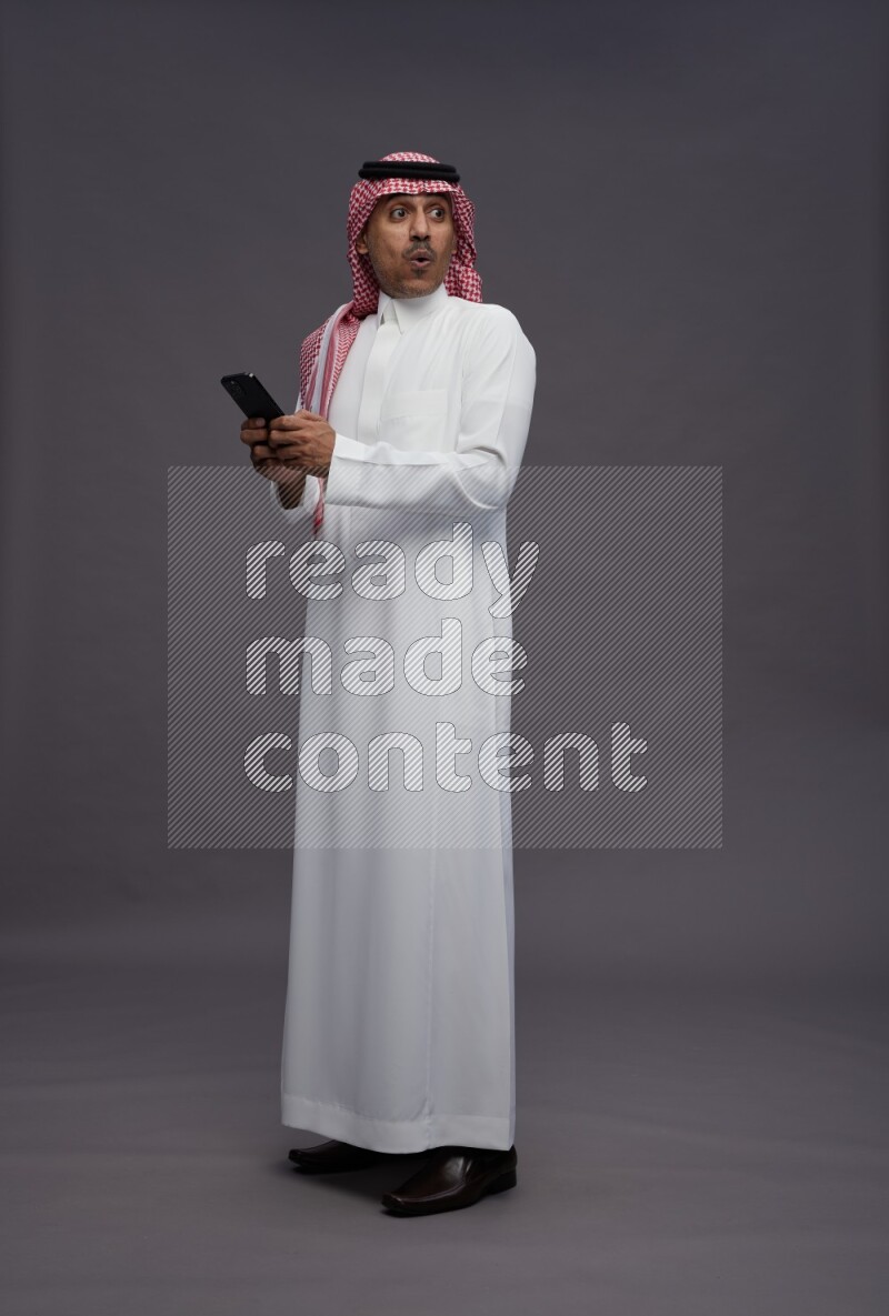 Saudi man wearing thob and shomag standing texting on phone on gray background