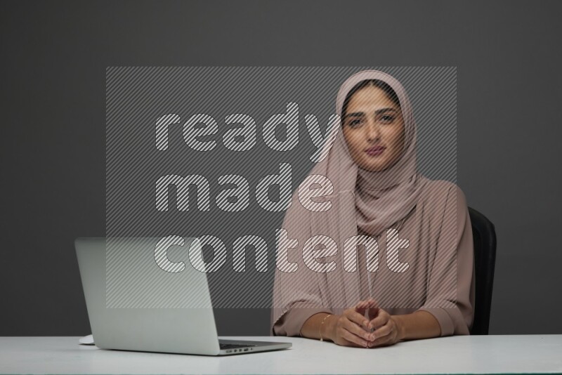 A Saudi woman Setting on her desk on a Gray Background wearing Brown Abaya with Hijab