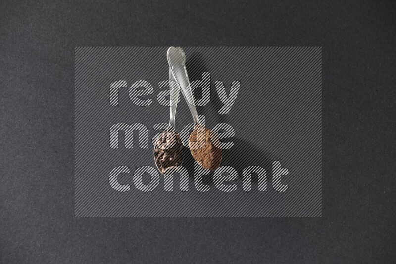 2 metal spoons, one full of cloves grains and the other full of cloves powder on a black flooring