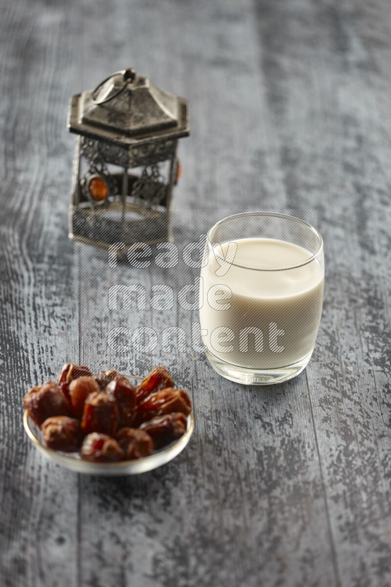A silver lantern with different drinks, dates, nuts, prayer beads and quran on grey wooden background