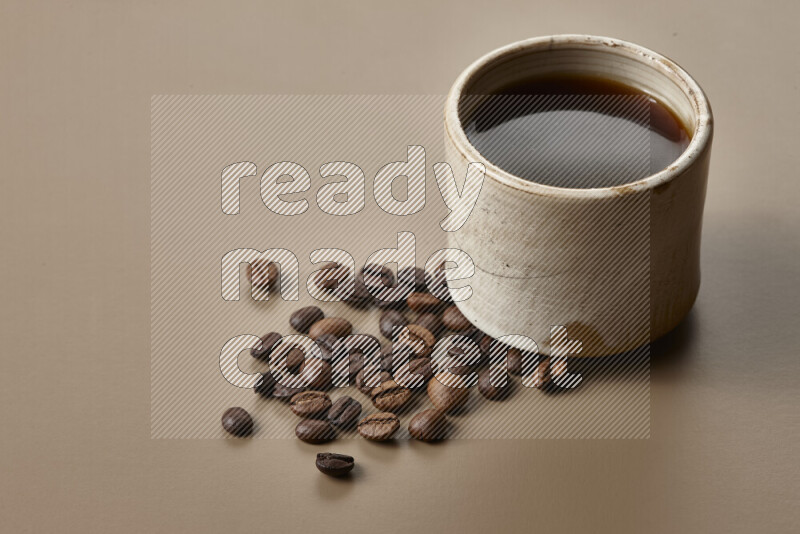 A beige pottery cup of coffee surrounded by roasted coffee beans on beige background