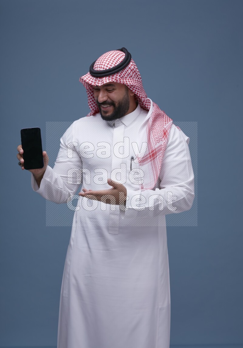 A man Showing his smartphone Screen on Blue Background wearing Saudi Thob