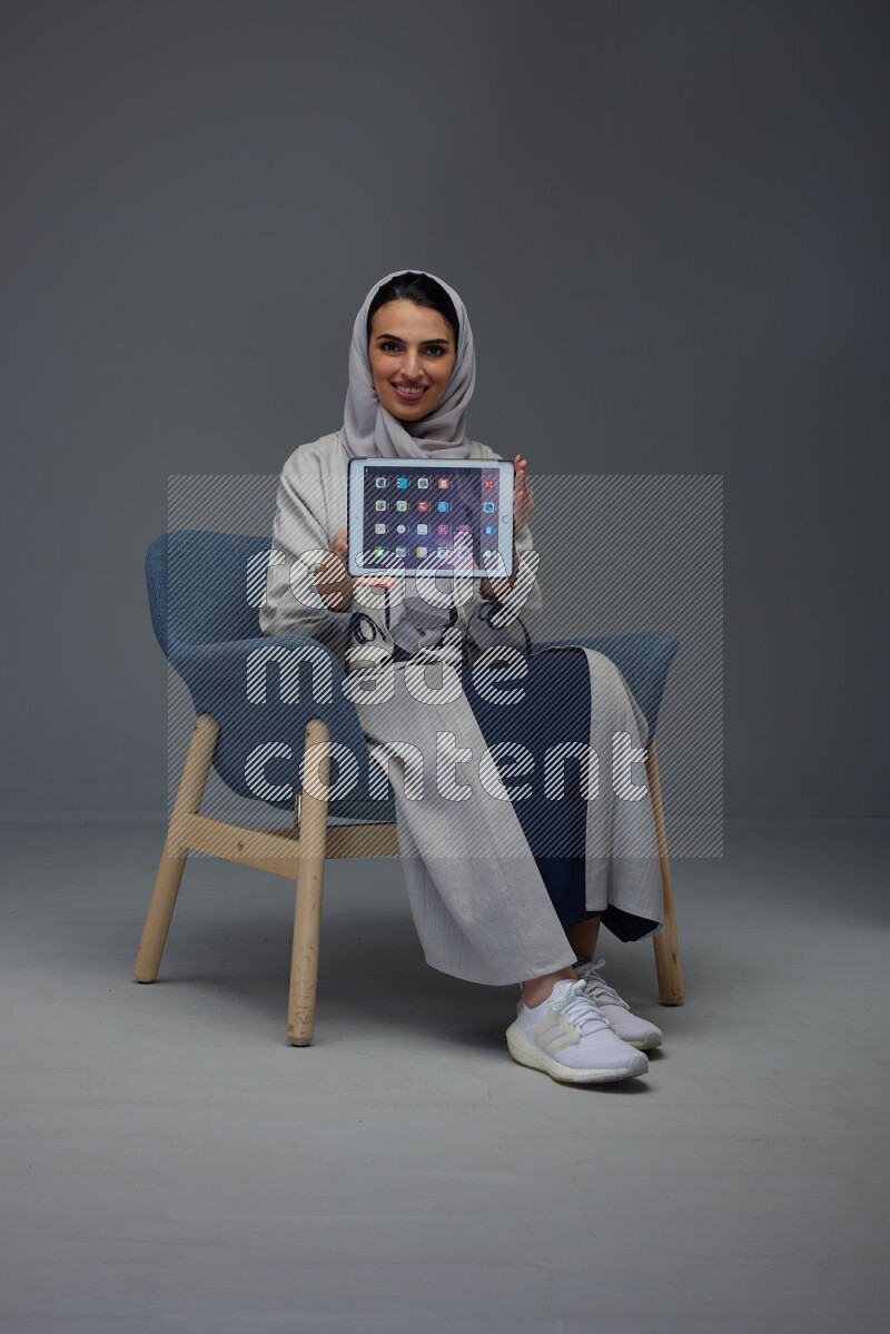 A Saudi woman wearing a light gray Abaya and head scarf sitting on a dark grey chair and showing the tablet's screen eye level on a grey background