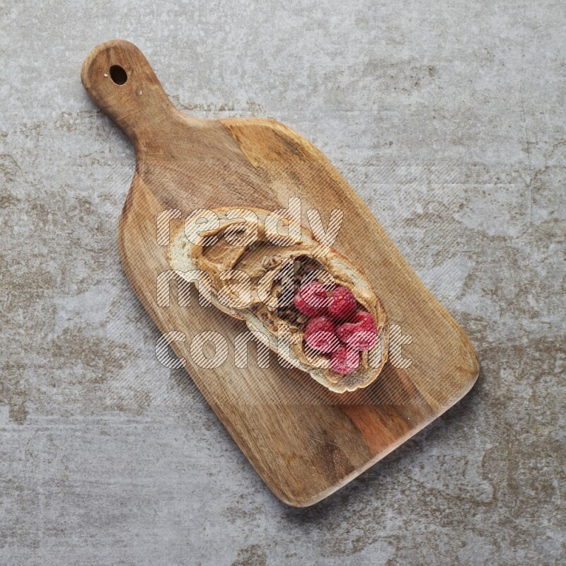 open faced peanut butter sandwich with raspberries and chocolate granula on a grey textured background