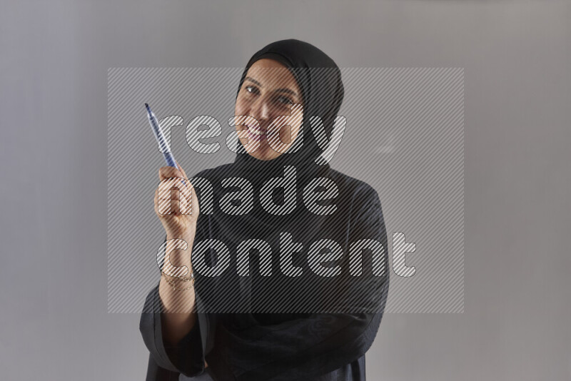 Woman in black abaya and hijab holding a marker pen to write on a transparent board with different reactions and expressions on grey background