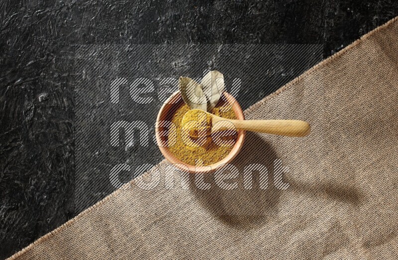 A wooden bowl and a wooden spoon full of turmeric powder on burlap fabric on textured black flooring