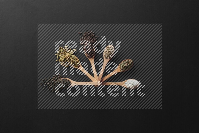 wooden spoons filled with white peppers, cloves, cardamom, salt, black peppers and basil on black flooring