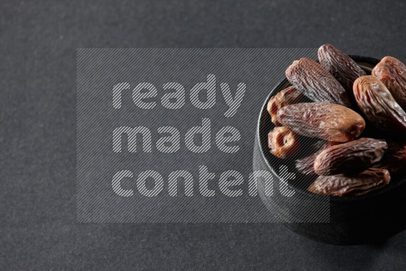 A black pottery bowl full of dried dates on a black background in different angles