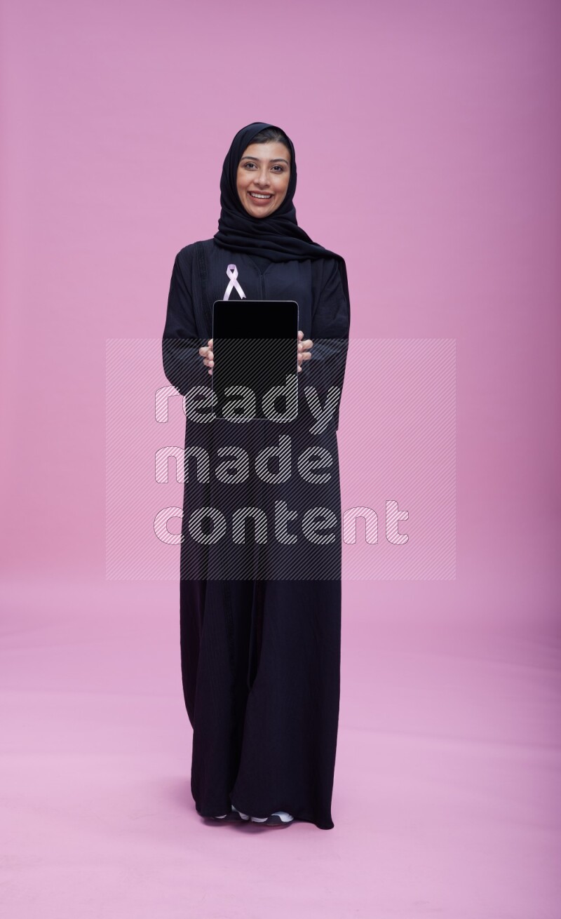 Saudi woman wearing pink ribbon on Abaya standing showing a tablet to camera on pink background