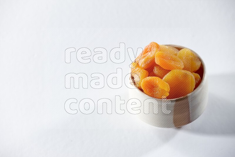 A beige ceramic bowl full of dried apricots on a white background in different angles