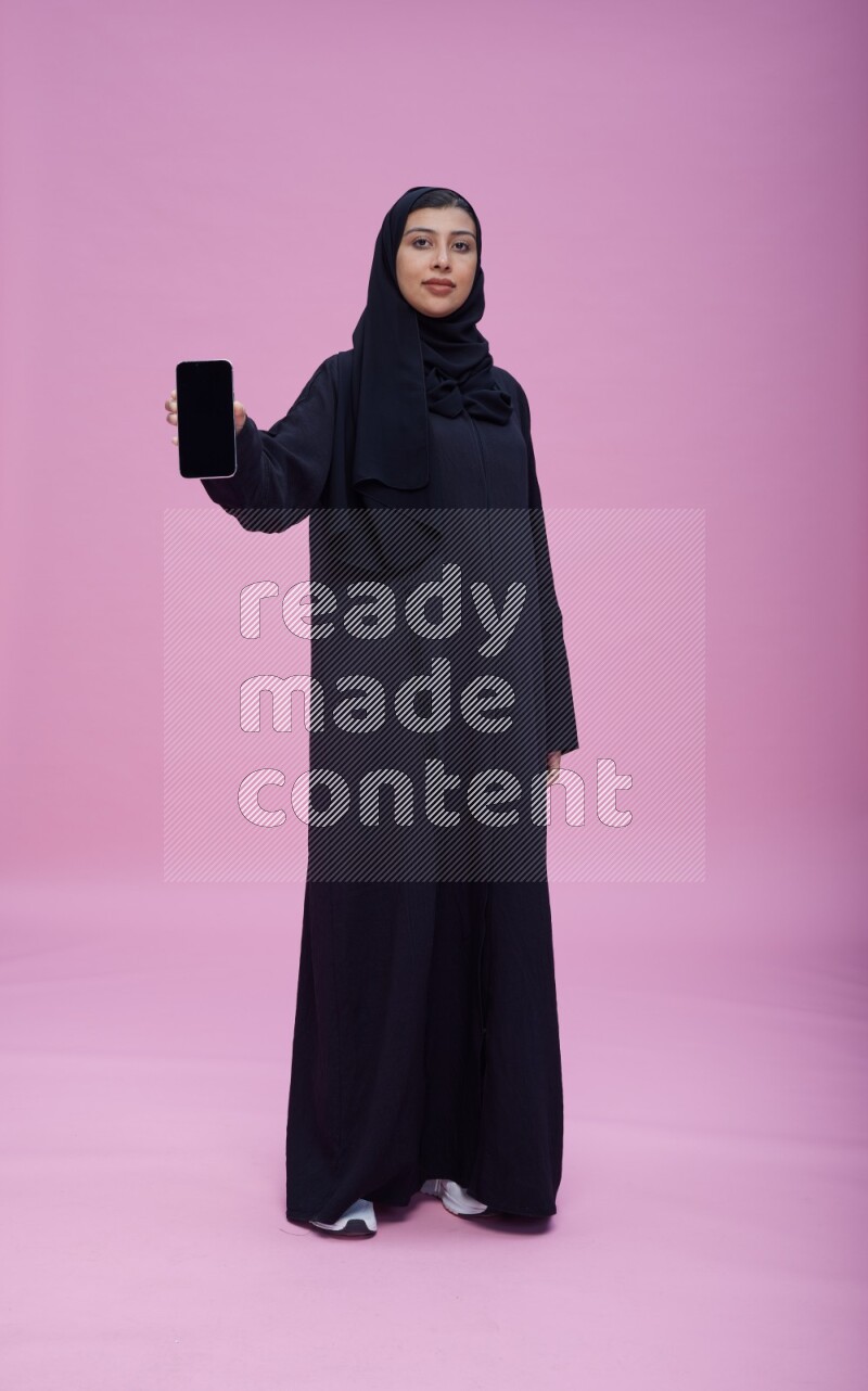 Saudi woman wearing Abaya standing showing phone to camera on pink background
