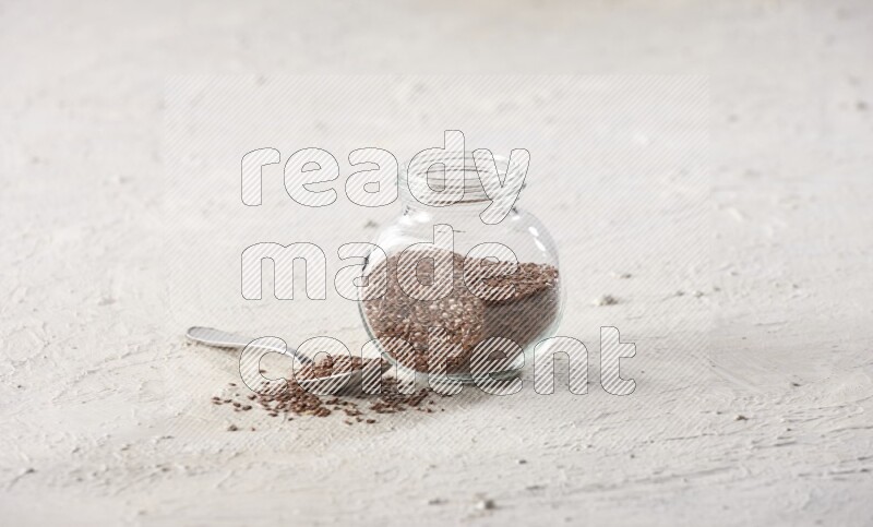 A glass spice jar full of flax seeds and a metal spoon full of the seeds on a textured white flooring