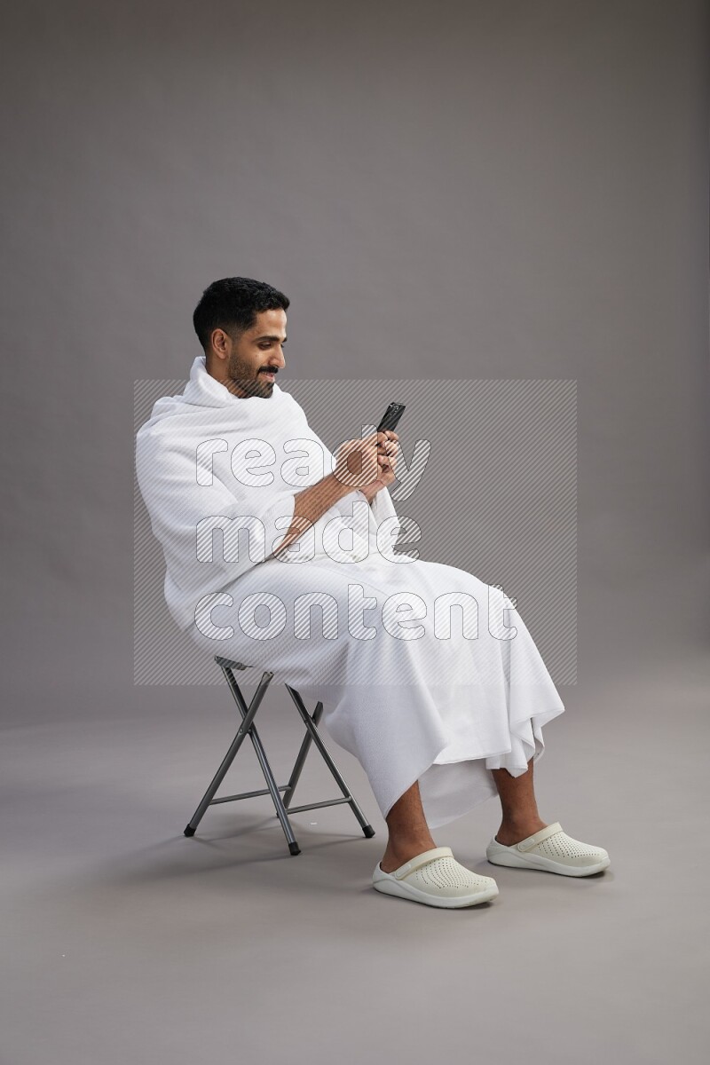 A man wearing Ehram sitting on chair texting on phone on gray background