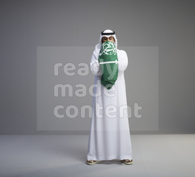 A Saudi man standing wearing thob and white shomag with face painting kissing big Saudi flag on gray background