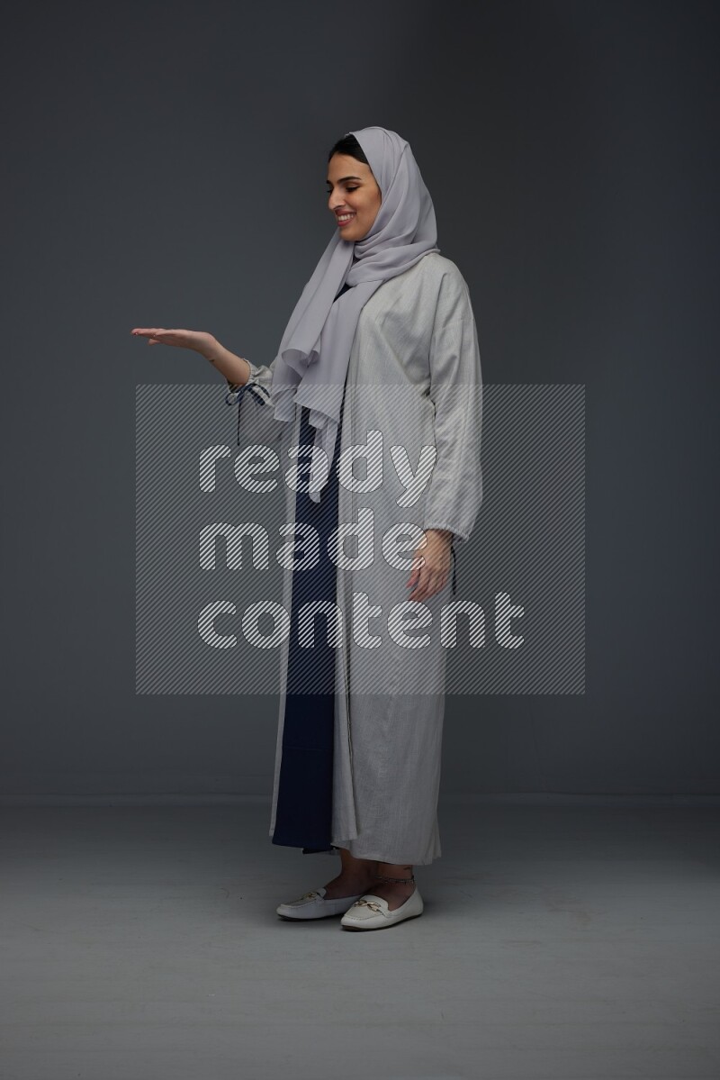 A Saudi woman wearing a light gray Abaya and head scarf standing and pointing in different directions eye level on a grey background