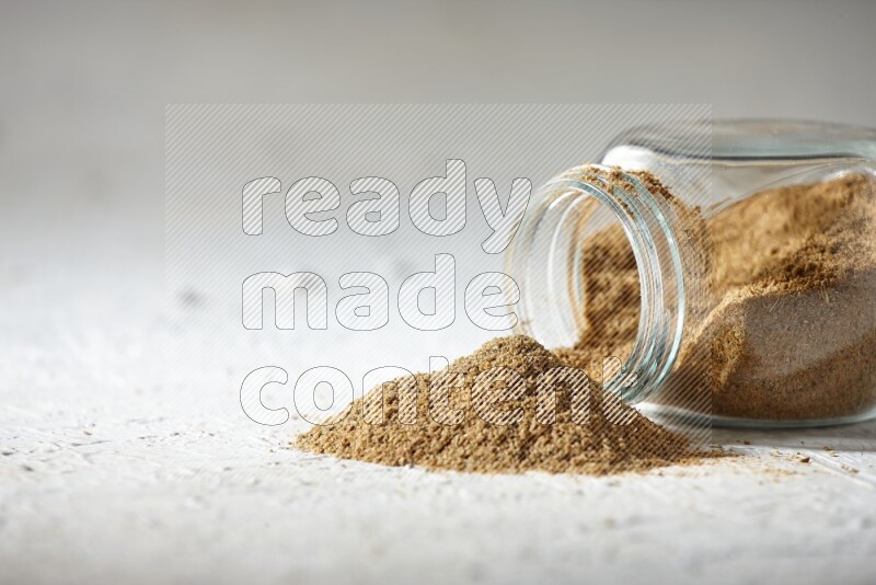A flipped glass spice jar full of cumin powder and powder spilled out on textured white flooring