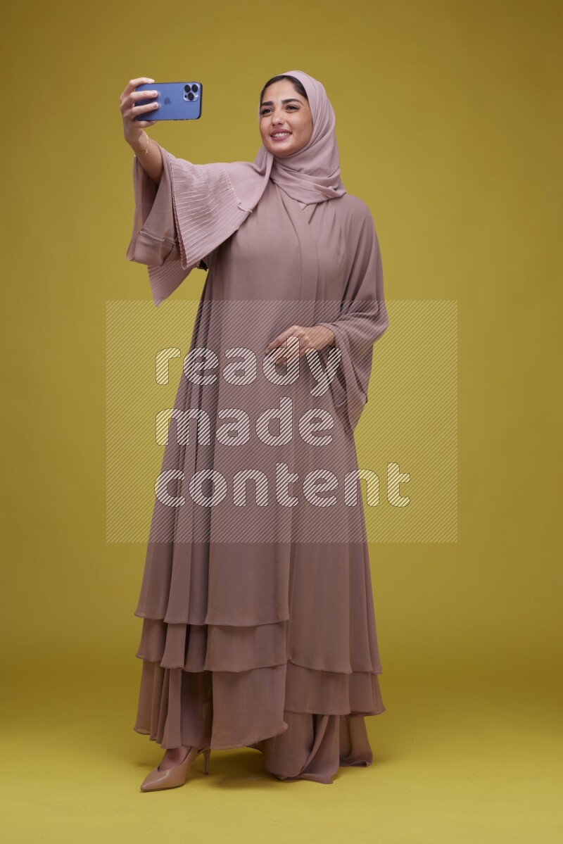 A woman taking a Selfie on a Yellow Background wearing Brown Abaya with Hijab