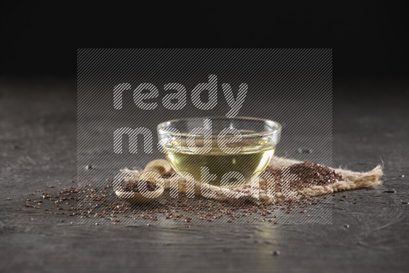 A glass bowl full of flaxseeds oil and wooden spoon full of flaxseeds with seeds spread on burlap fabric on a textured black flooring