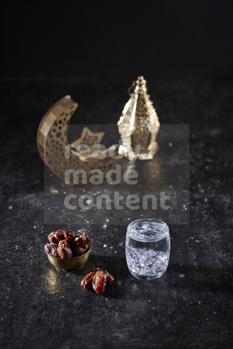 Dates in a metal bowl with water beside golden lanterns in a dark setup