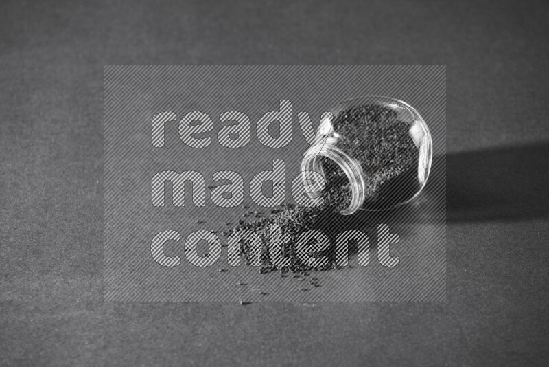 A flipped glass spice jar full of black seeds and the seeds spread out on a black flooring