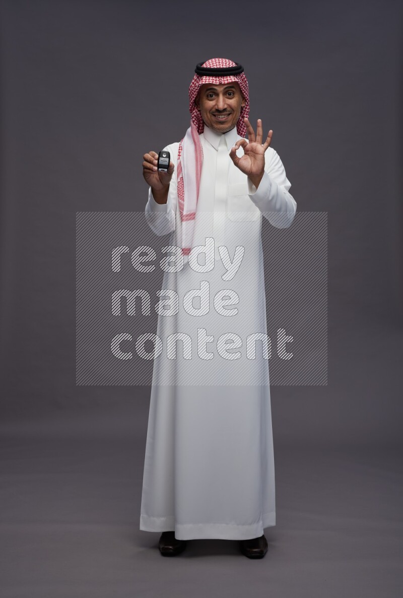 Saudi man wearing thob and shomag standing holding key car on gray background