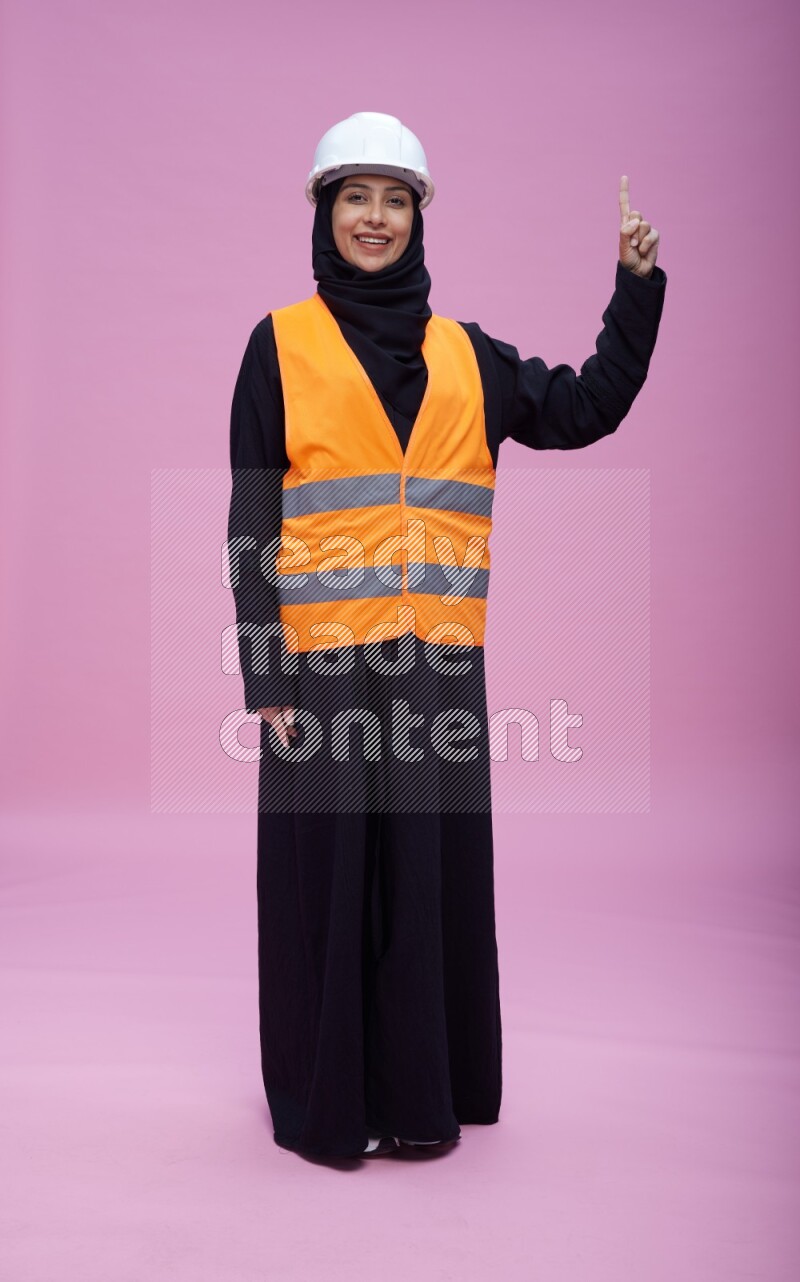 Saudi woman wearing Abaya with engineer vest and helmet standing interacting with the camera on pink background