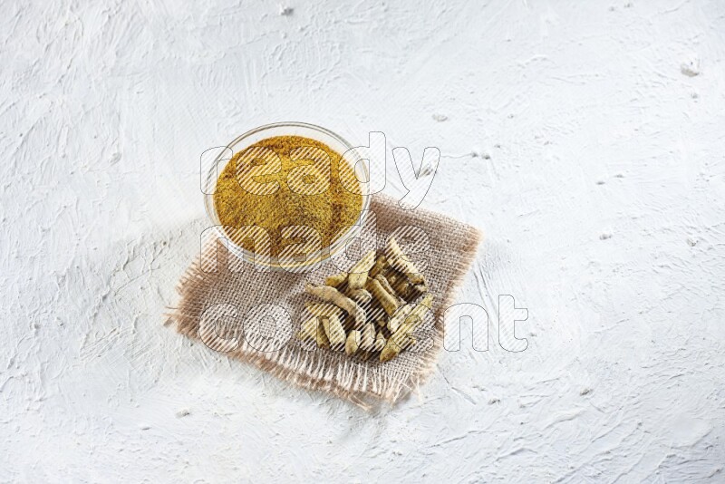A glass bowl full of turmeric powder and dried turmeric whole finger on a piece of burlap on a textured white flooring
