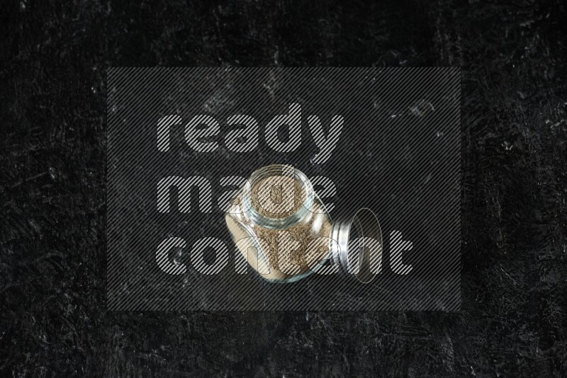 A glass spice jar full of cardamom powder on textured black flooring