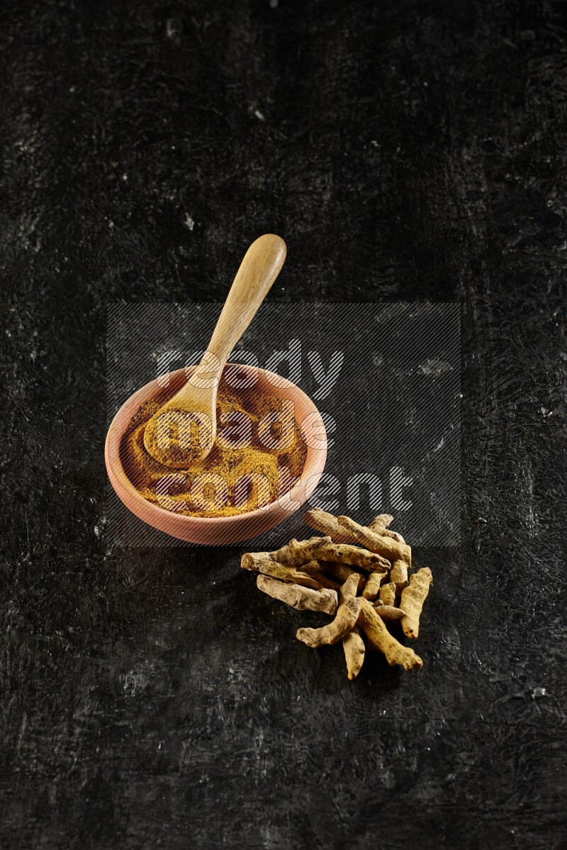 A wooden bowl and a wooden spoon full of turmeric powder with dried turmeric fingers on textured black flooring