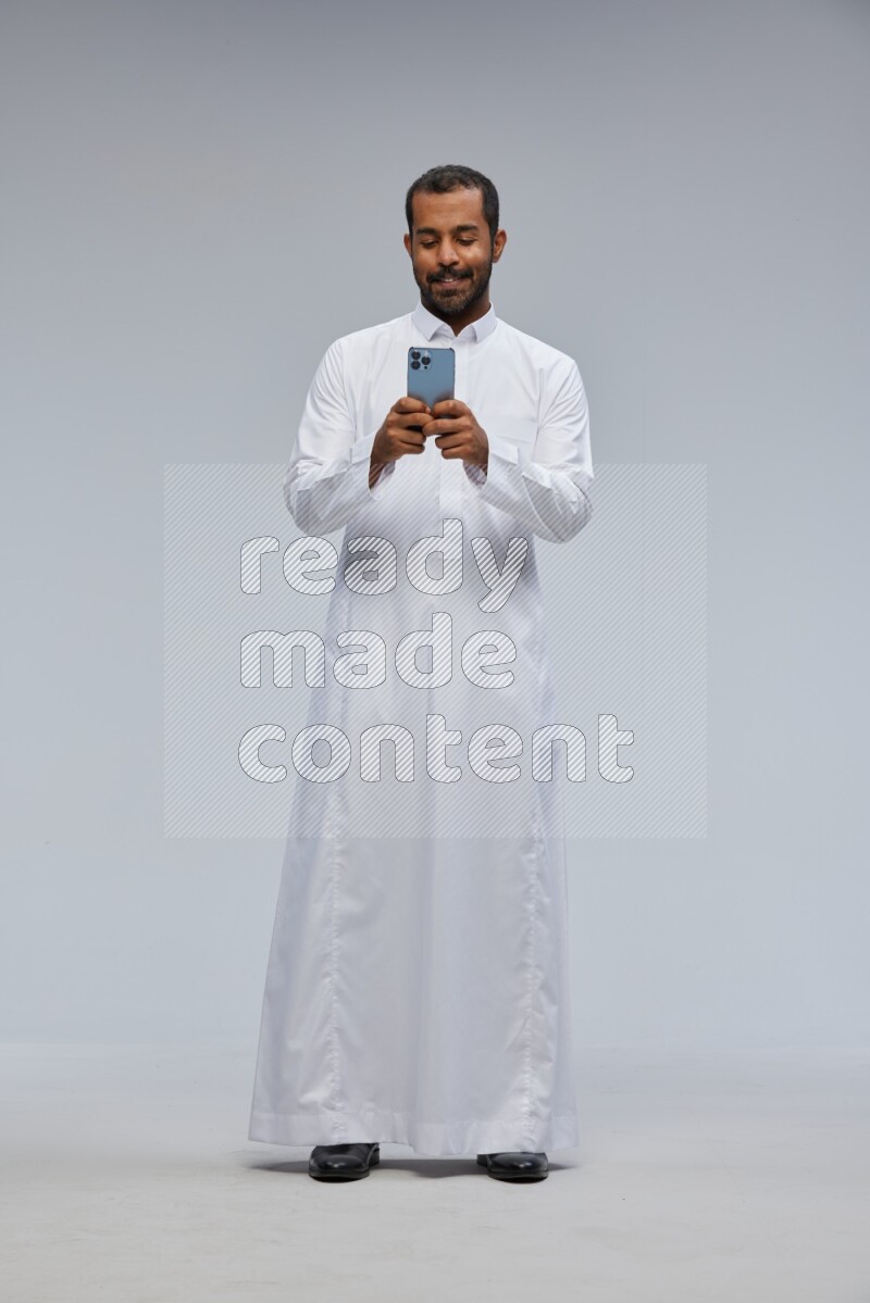 Saudi man wearing Thob standing texting on phone on Gray background