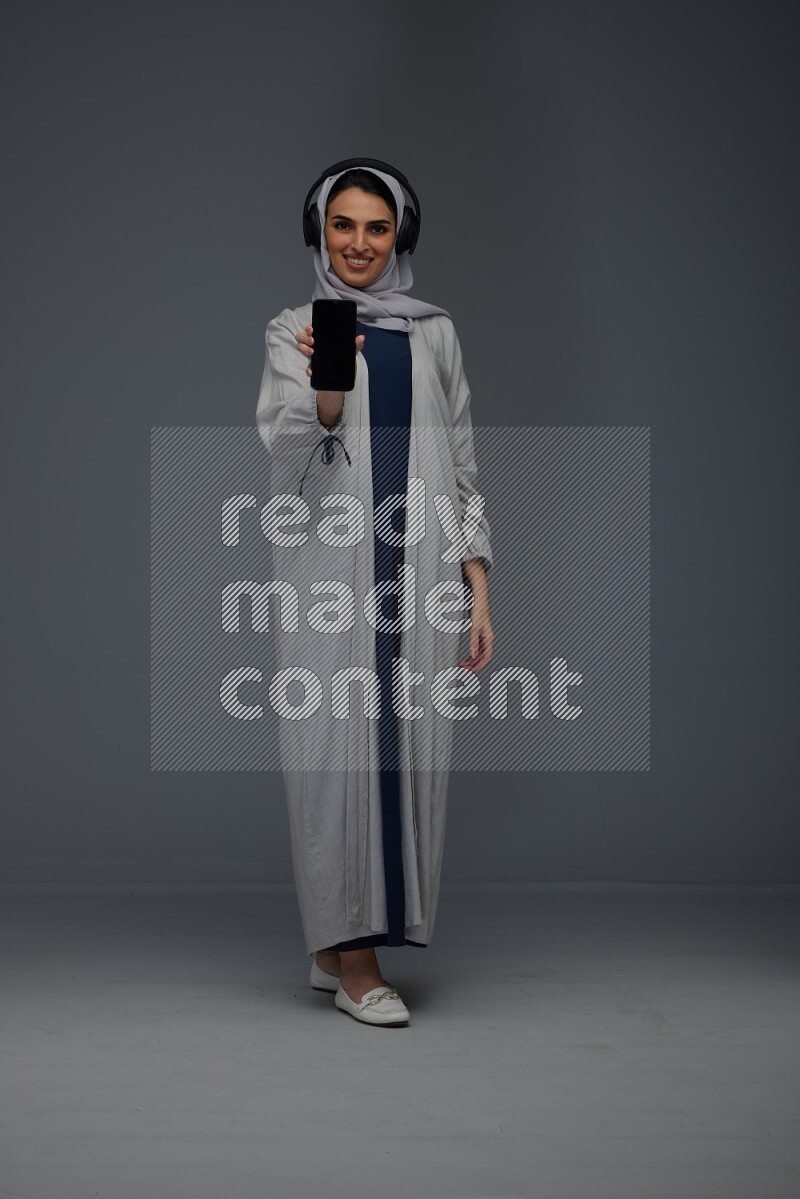 A Saudi woman wearing a light gray Abaya and head scarf setting on a dark grey chair and watching on the tablet eye level on a grey background