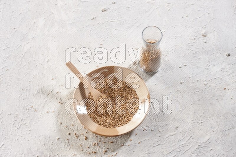 A beige pottery plate full of mustard seeds and a wooden spoon in it with a glass jar filled with the seeds on a textured white flooring