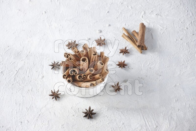 White bowl full of cinnamon sticks surrounded by star anis on a textured white background in different angles