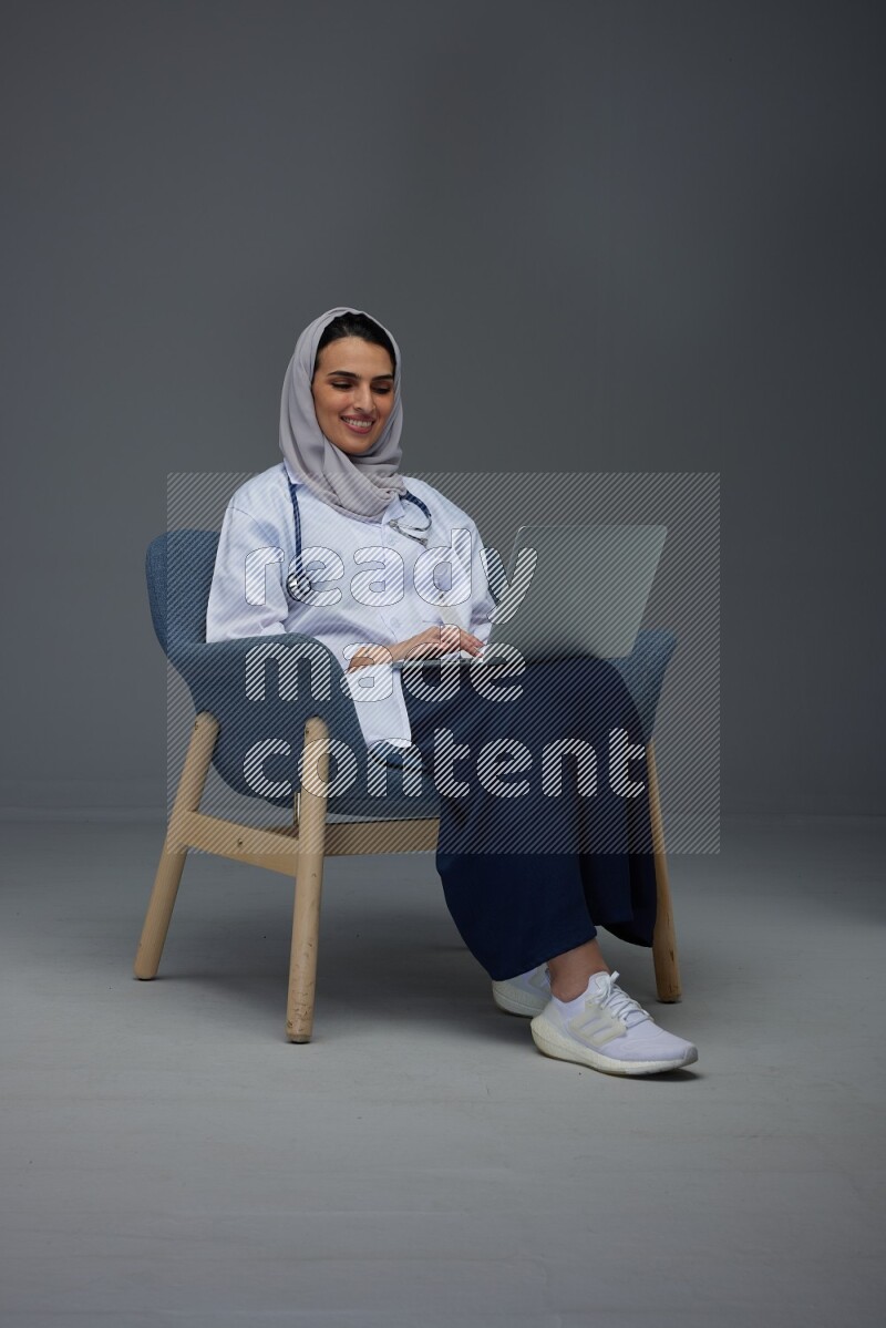 A doctor wearing a light gray head scarf sitting on a dark grey chair and using a laptop eye level on a grey background