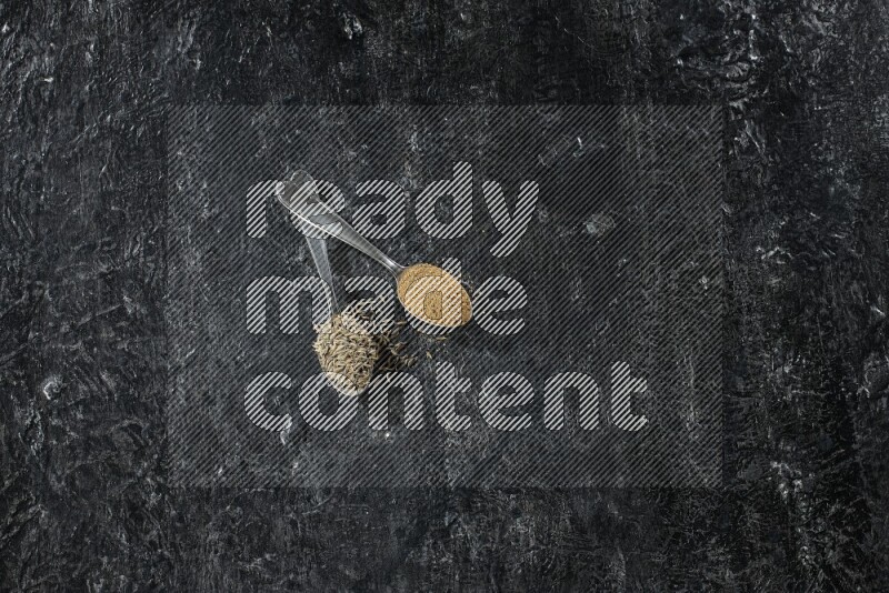 2 metal spoons full of cumin seeds and cumin powder on a textured black flooring