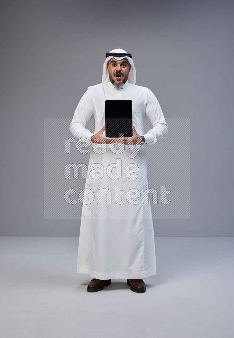 Saudi man Wearing Thob and white Shomag standing showing tablet to camera on Gray background
