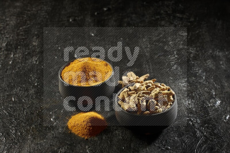 2 pottery black bowls, one full of turmeric powder and the other full of dried turmeric whole fingers on a textured black flooring