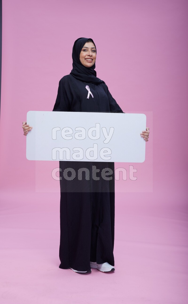 Saudi woman wearing pink ribbon on Abaya standing holding board on pink background