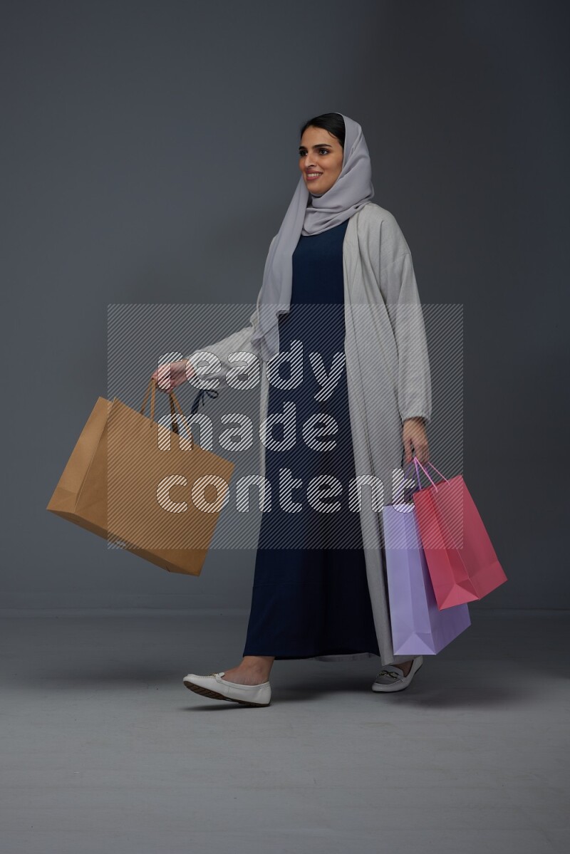 A Saudi woman wearing a light gray Abaya and head scarf standing and holding shopping bags on a grey background